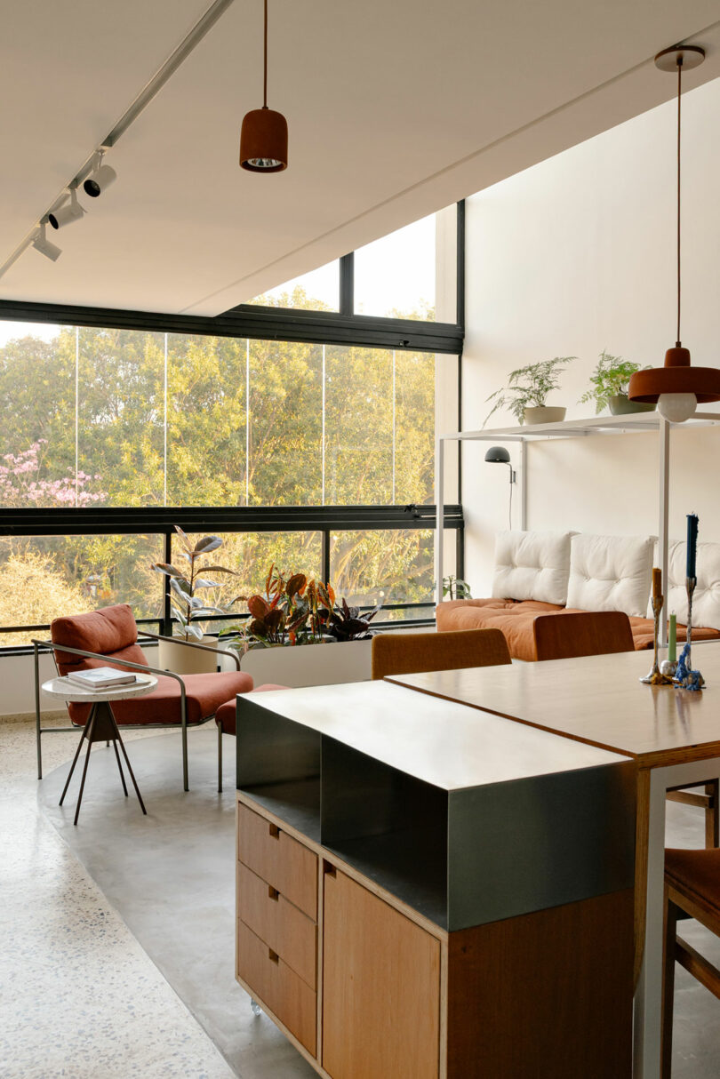 Living space with orange chairs and large windows showing lush green trees outside.