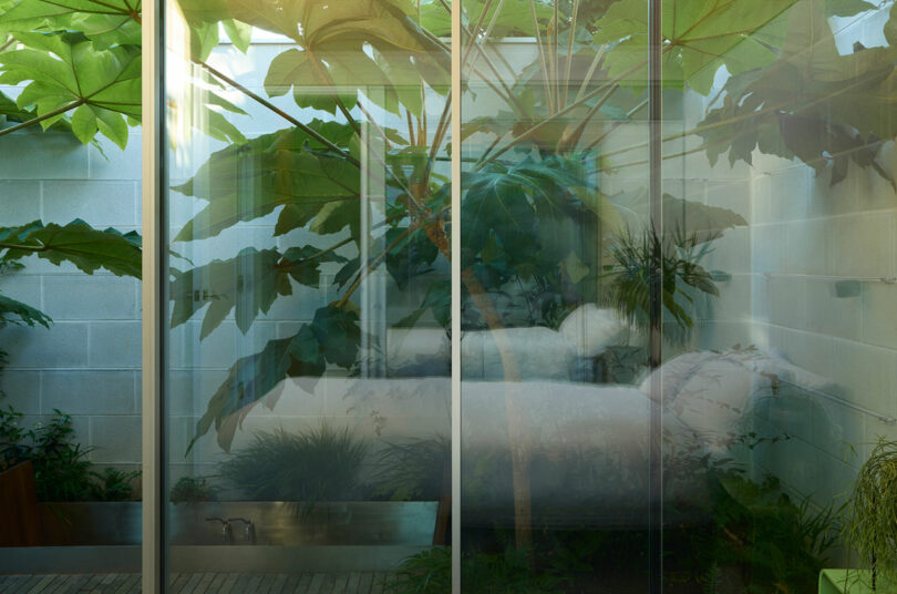 A minimalist bedroom with white bedding separated from a dense green garden by a large floor-to-ceiling glass wall.