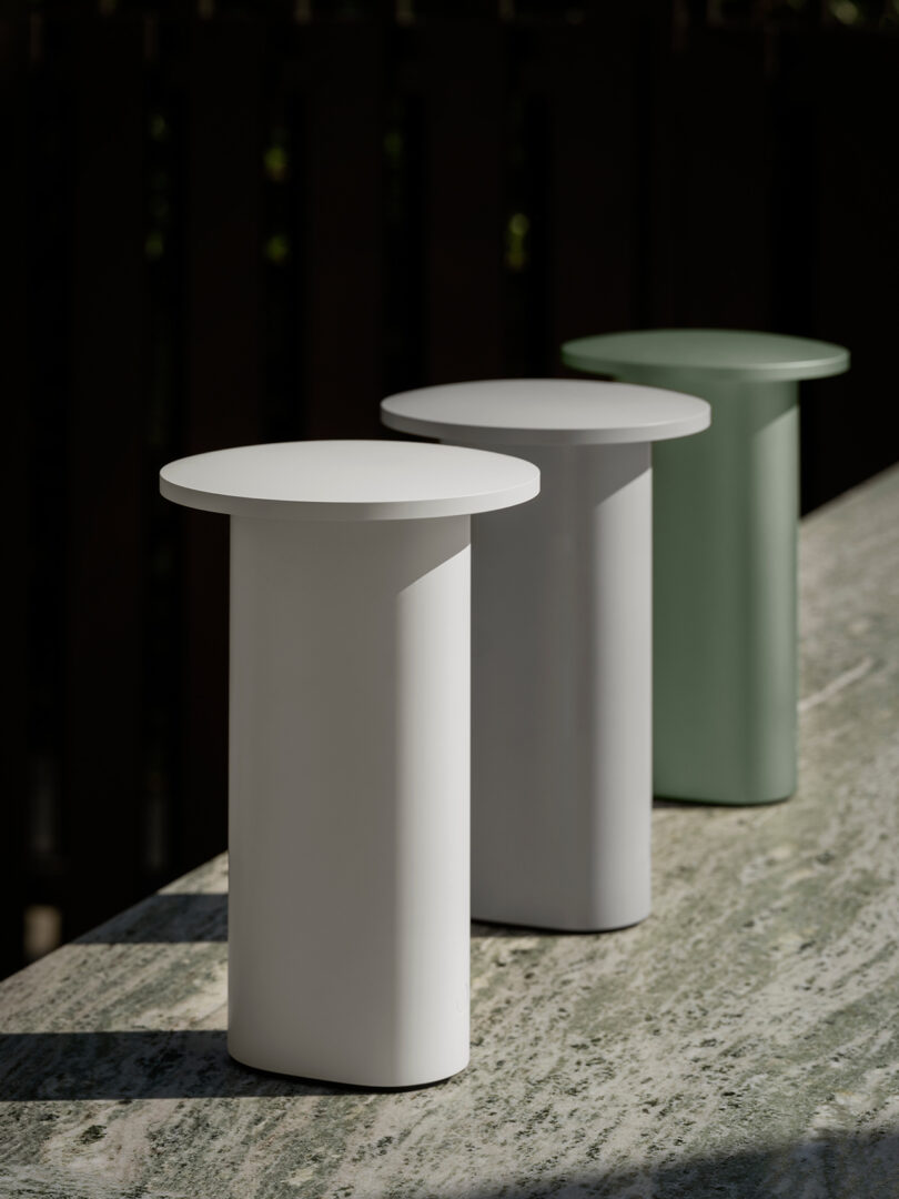 Three cylindrical side tables or lamp bases in white, gray, and green arranged on a marble surface.