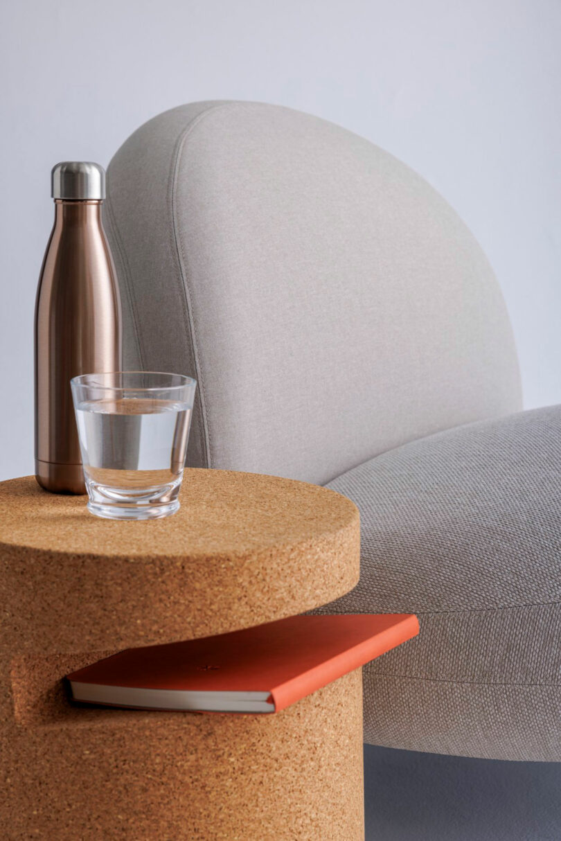 A round cork table next to a lounge chair holding a water bottle, a glass, and a notebook.
