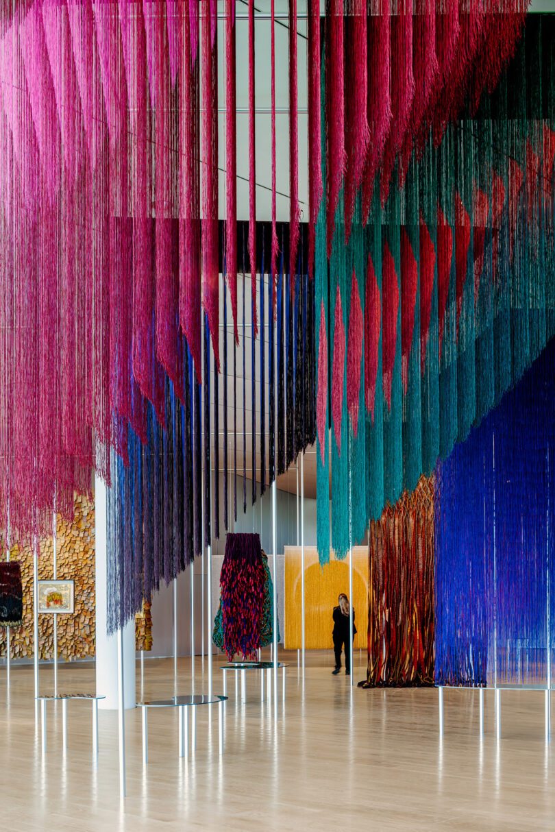 Vibrant hanging thread installations in shades of blue, pink, and orange in a gallery.