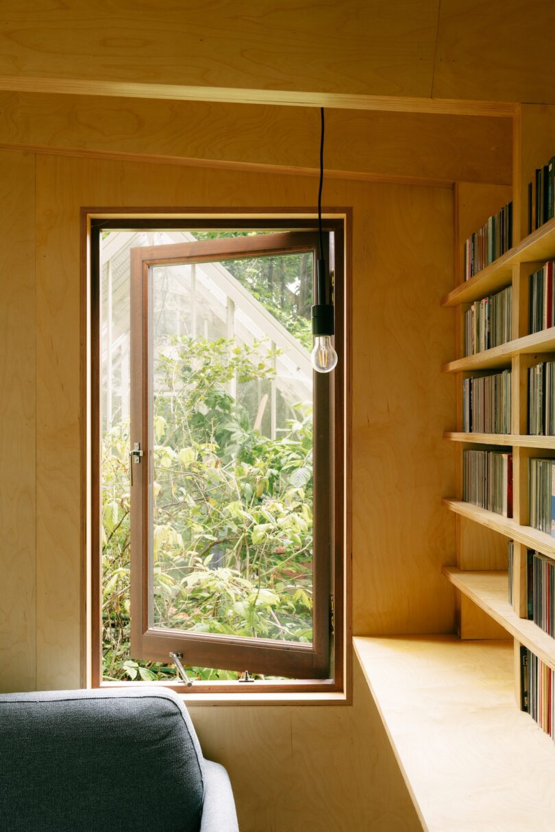 A cozy interior corner with a pendant light, a gray sofa, and a wall of music media next to a window view of greenery.