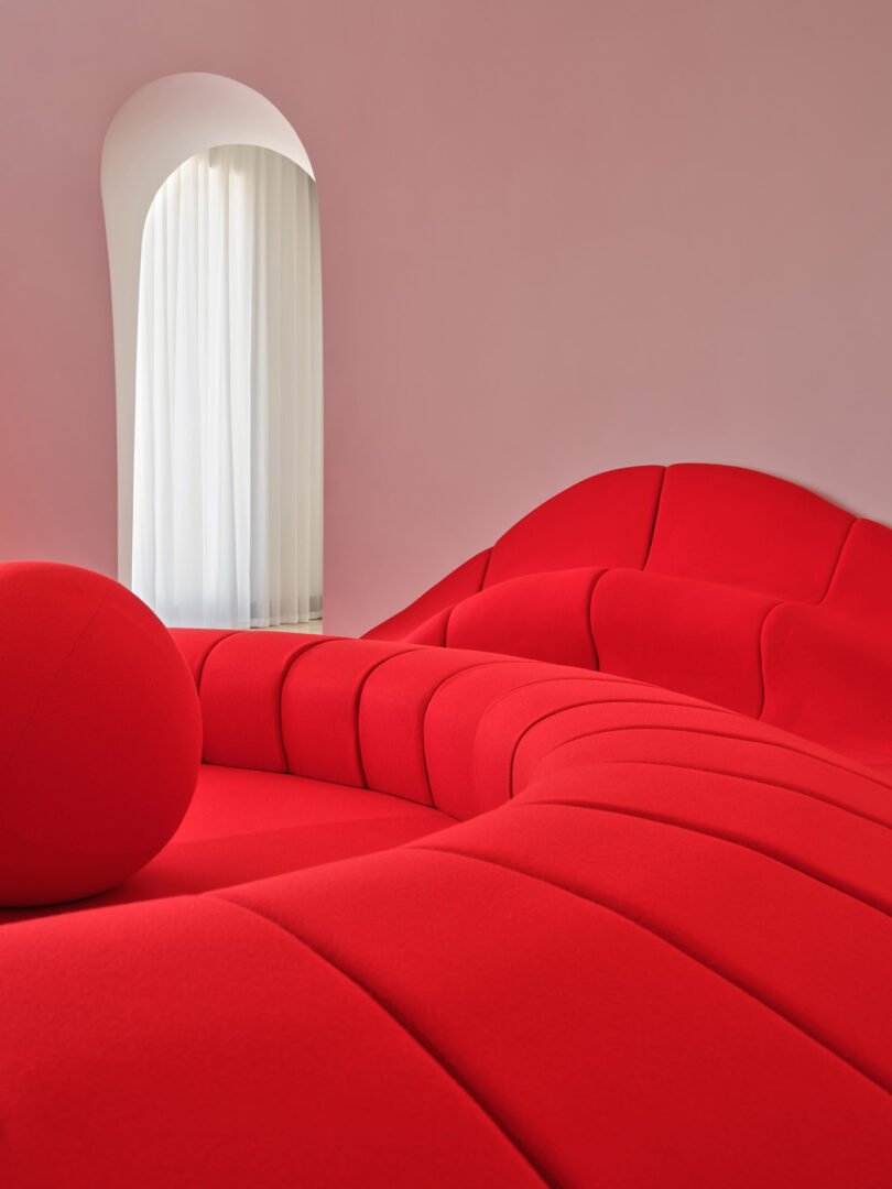 A bright red sculptural sofa set against a pale pink wall with an arched window.