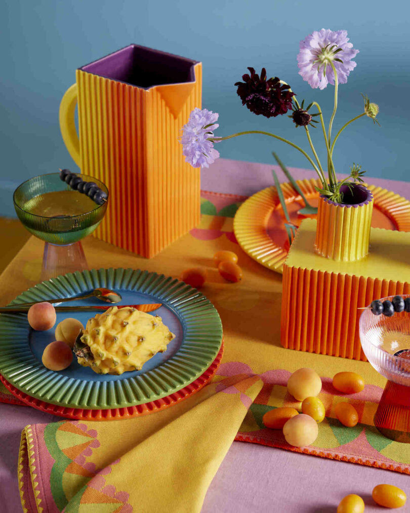 A curated table setting with a pitcher, vase, and plates on a patterned tablecloth.