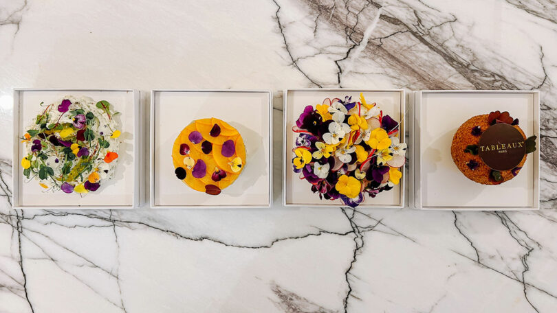 Artisanal desserts with floral toppings organized in white square boxes on a marble surface.