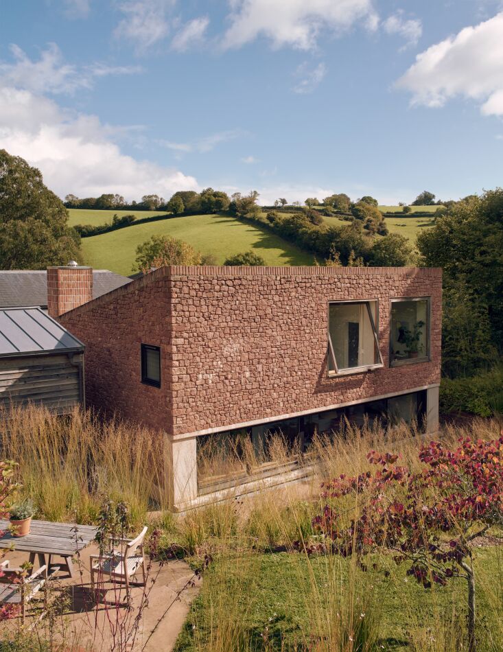 Exterior view of a pink sandstone house with reclaimed brick chimneys overlooking a vast meadow.