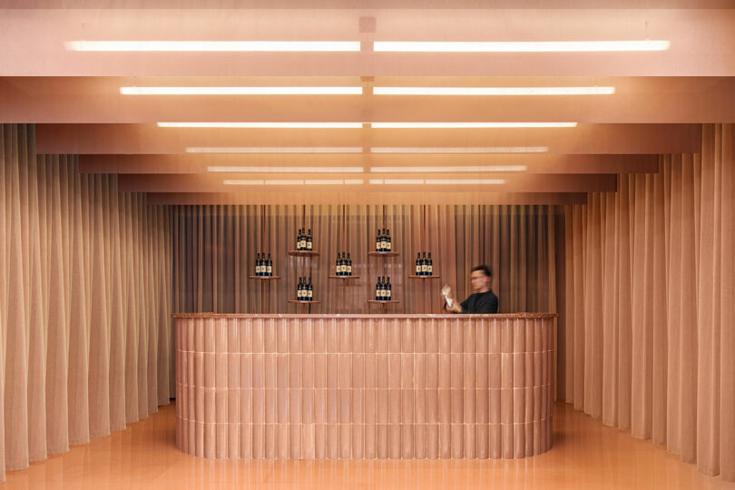 A bartender behind a minimalist curved bar with warm lighting and beige curtains.