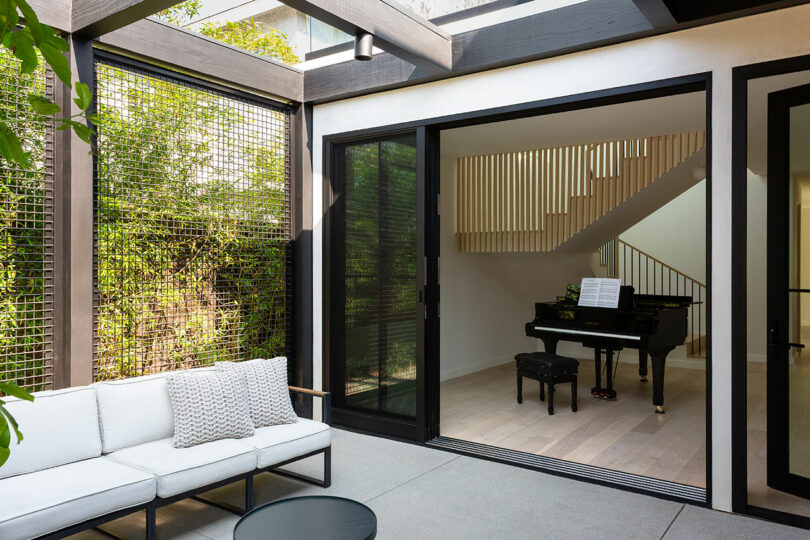 A modern living area featuring a black grand piano and a staircase with vertical wooden slats next to an outdoor sofa.