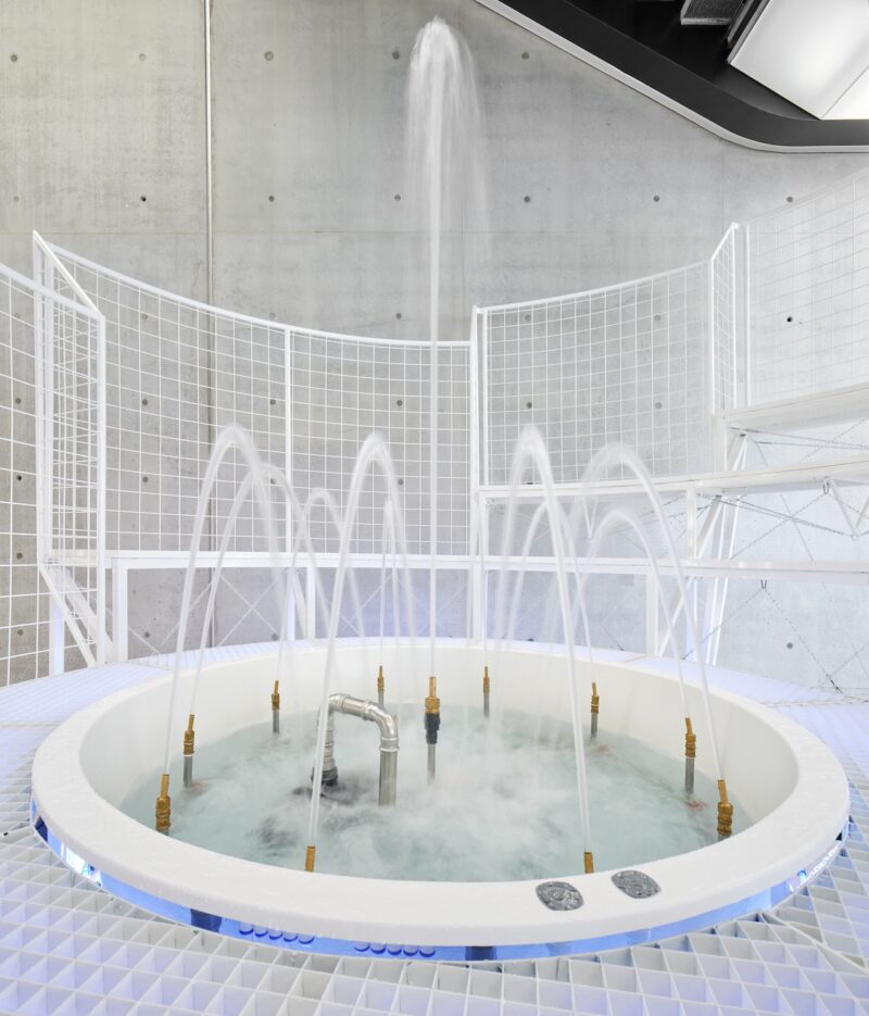 A circular indoor fountain with multiple spray heads surrounded by a white metal railing in a modern gallery.