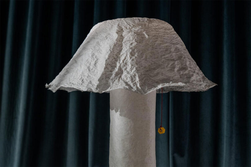 A floor lamp with a crumpled, textured white paper shade standing next to dark pleated curtains.
