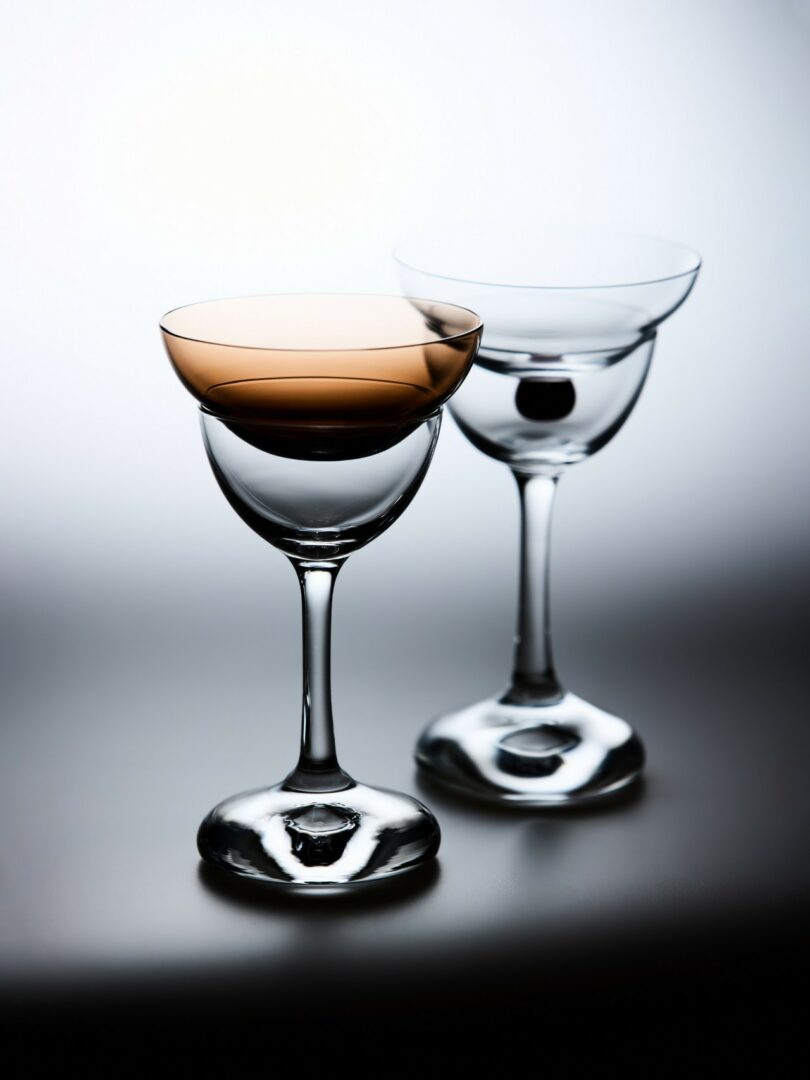 Two empty coupe glasses, one tinted brown and one clear, reflecting light on a polished surface.