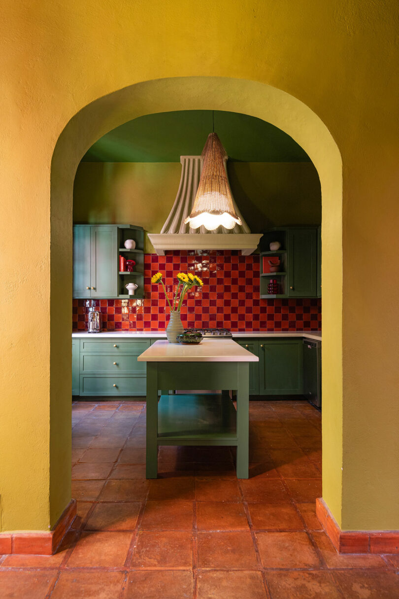 A kitchen viewed through an arched doorway, featuring green cabinets and a red checkered tile backsplash.