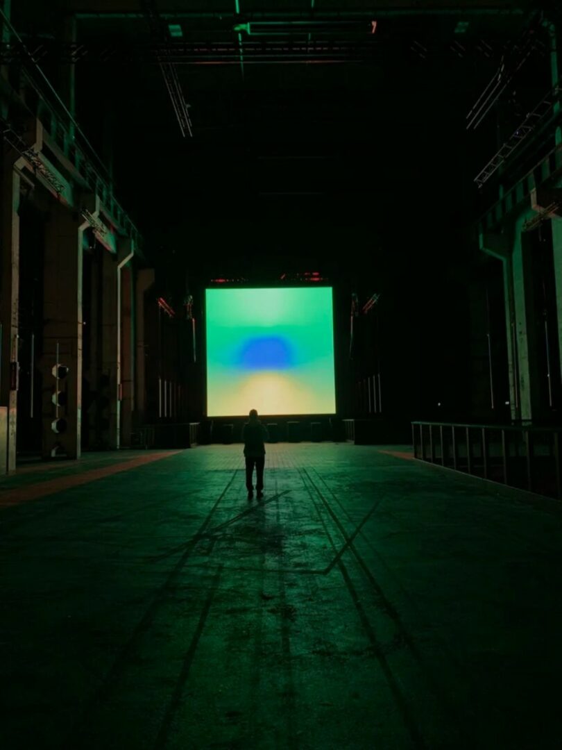 A lone silhouette facing a glowing green and blue abstract digital screen in a dark industrial room.