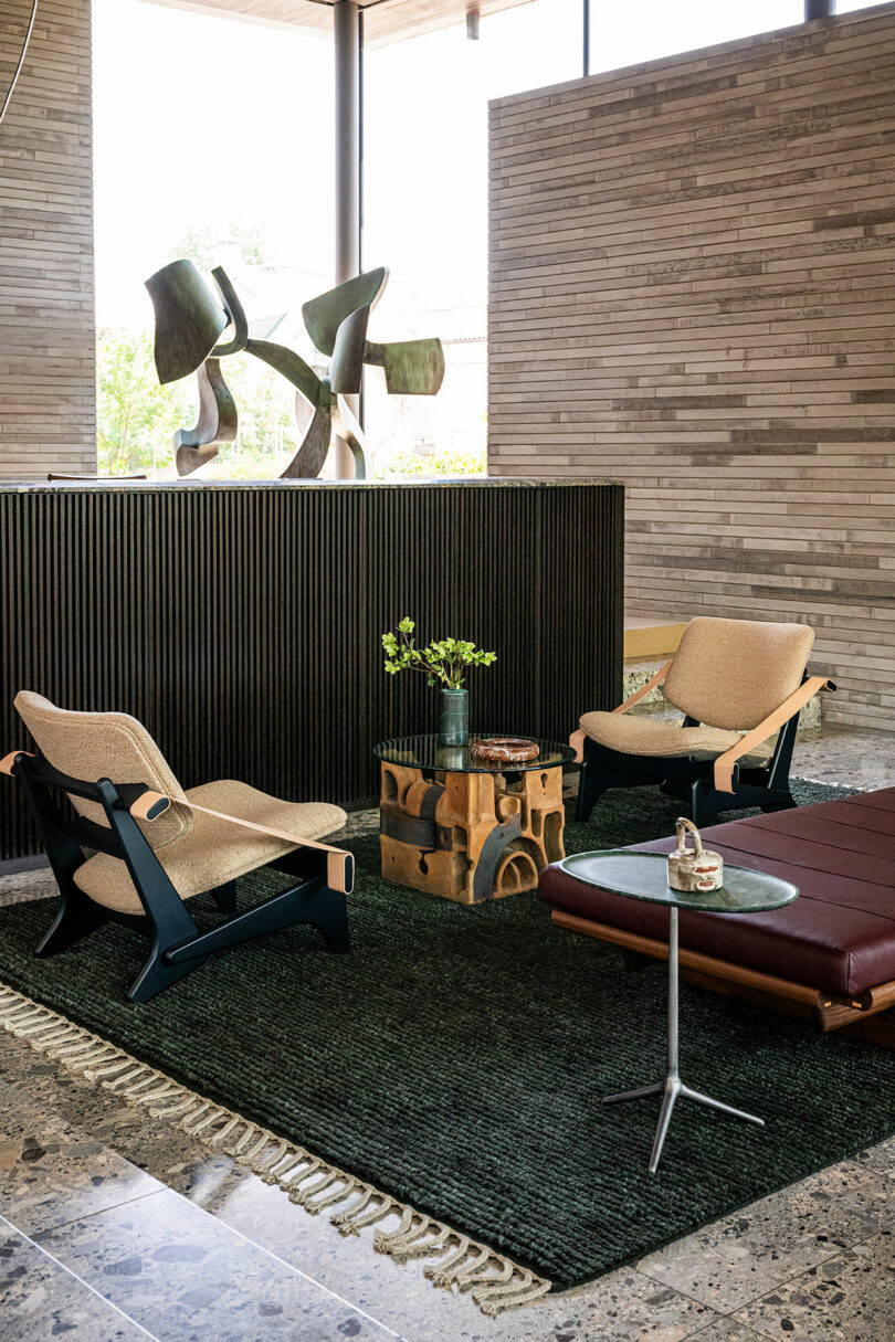 A modern lounge area with two armchairs, a leather bench, and a sculptural wooden coffee table on a stone floor.