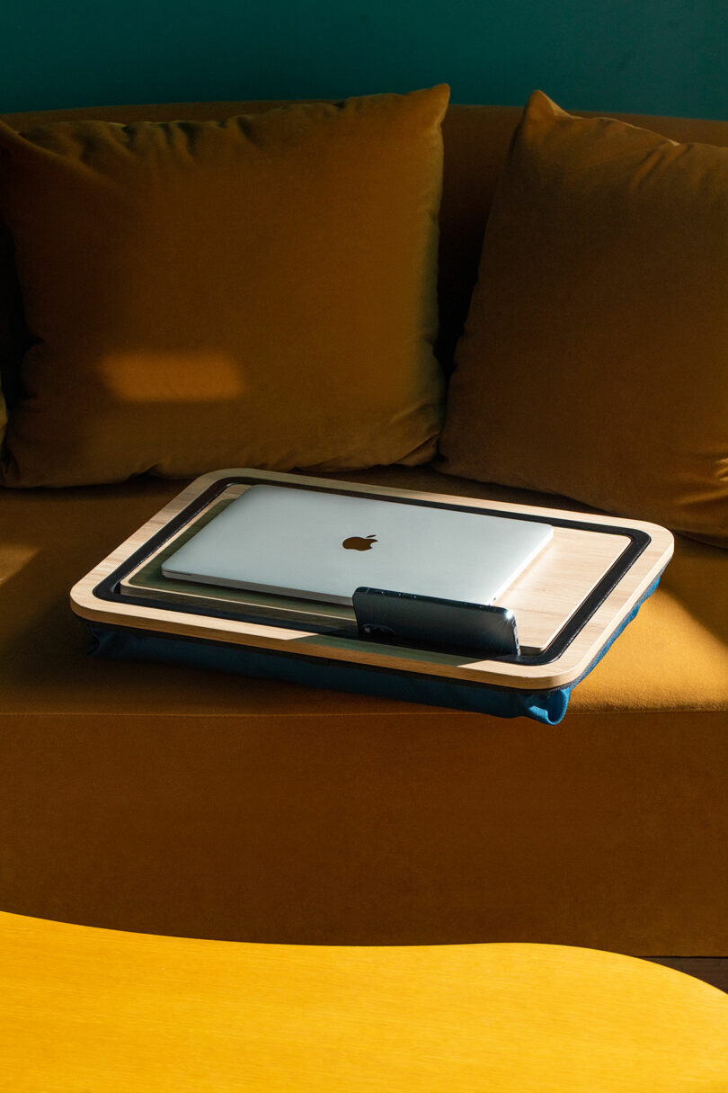 A closed MacBook and smartphone on a wooden lap desk resting on a yellow sofa.
