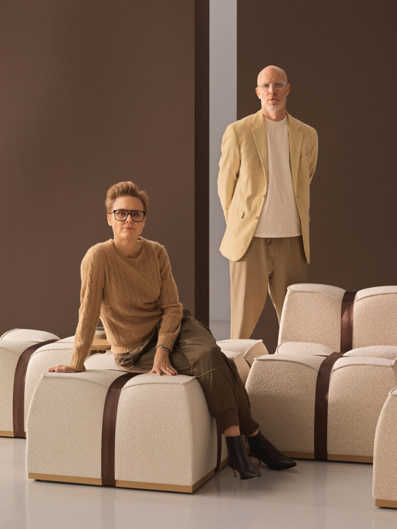 Two people in neutral clothing posing with cream-colored modular seating blocks against a brown background.