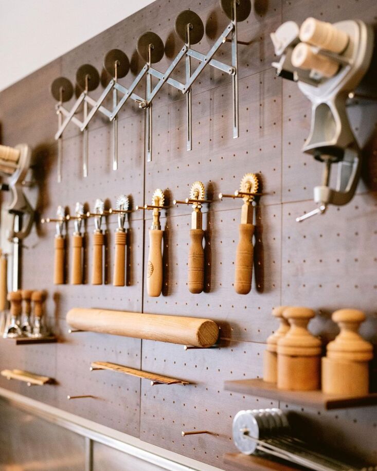 Close-up of kitchen utensils hanging from brass nails on a pegboard wall.