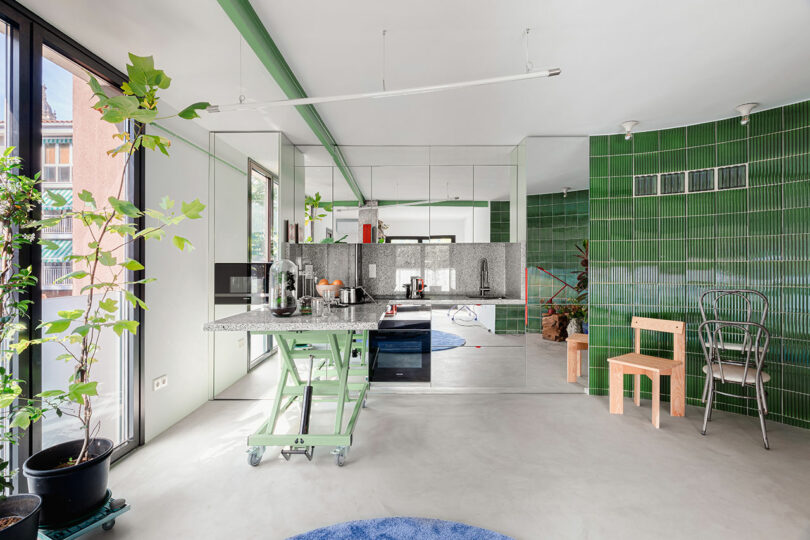 A modern kitchen area with an adjustable-height table and diverse chairs set against a green tiled backdrop.