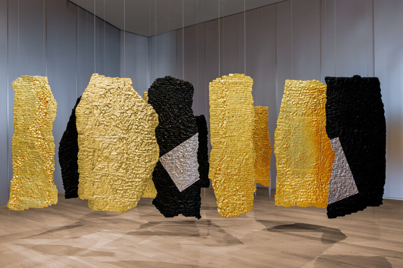 Large textured panels in gold, black, and silver suspended in a minimalist gallery.