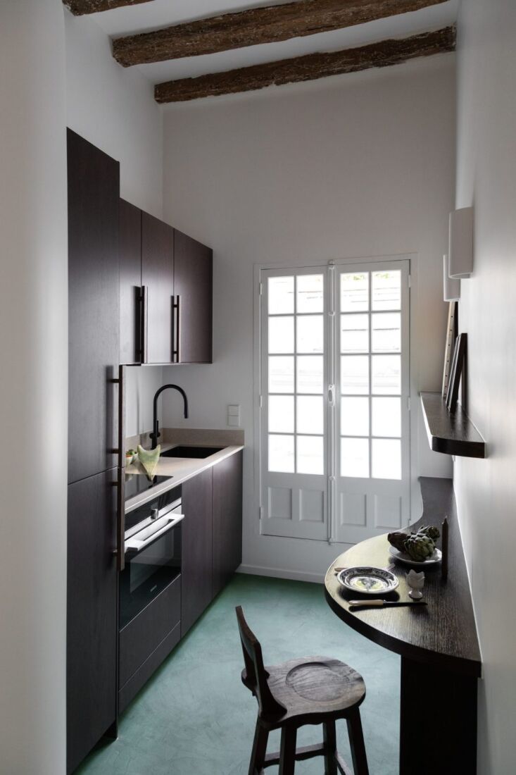 A kitchen with dark-stained oak cabinets and green-colored concrete flooring transition to oak planks.