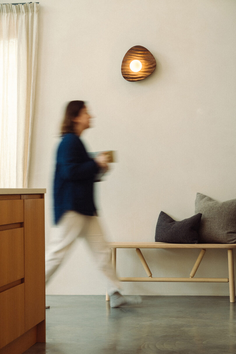 A person walks through a minimalist room with a round wooden wall light and a simple bench.