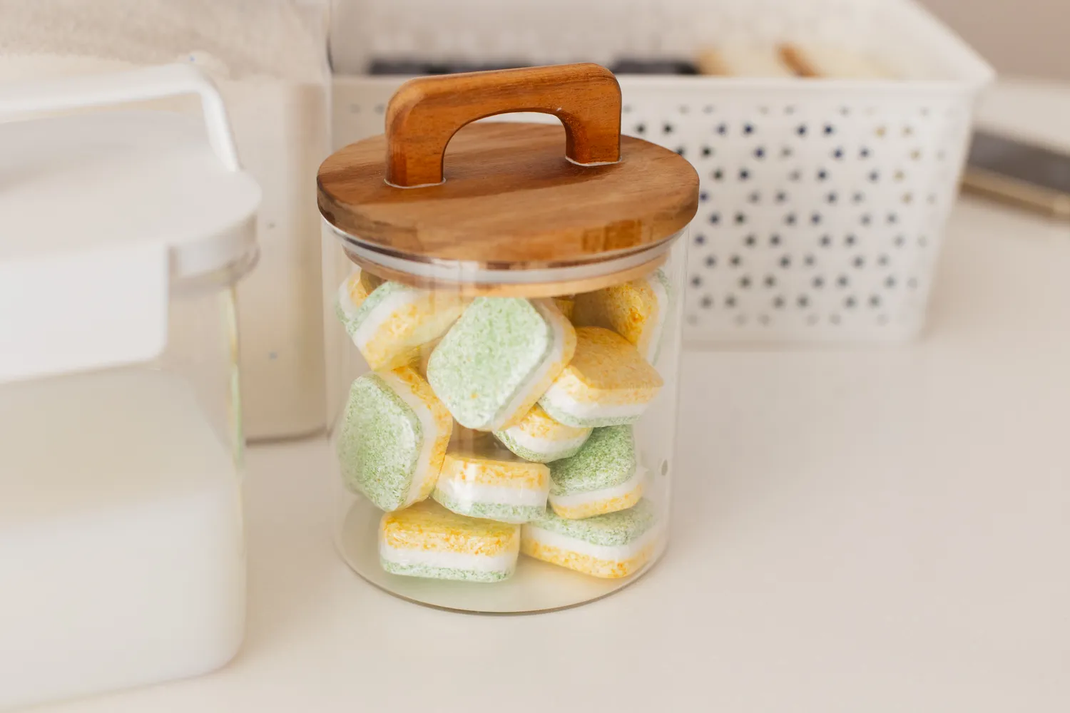A glass jar filled with multi-colored dishwasher tablets on a bright kitchen countertop.