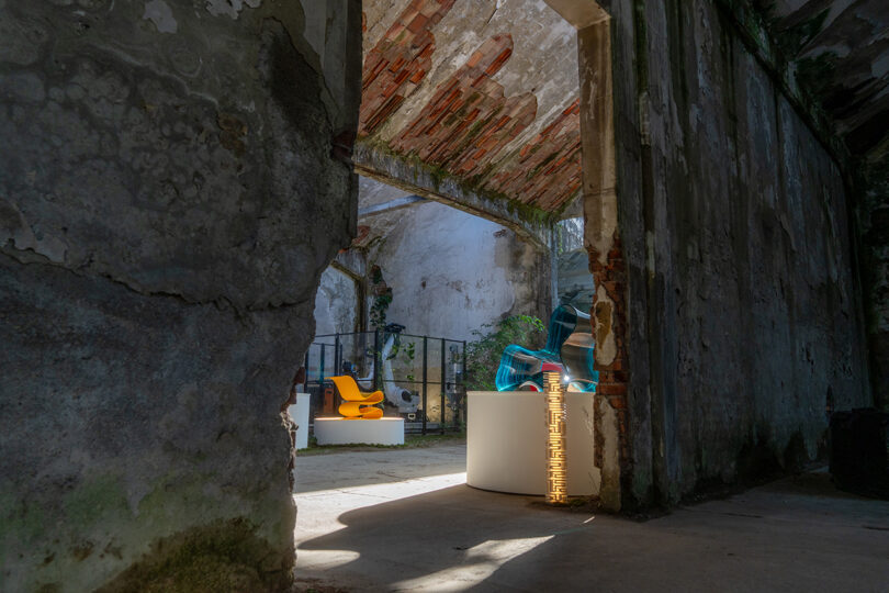 A view through a broken wall into a sunlit room filled with modern 3D-printed chairs.
