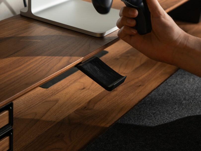 A hand attaching a black metal controller holder to the underside of the wooden desk shelf.