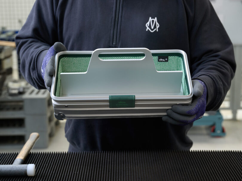 A person holding the RIMOWA x Vitra aluminum toolbox with green accents in a workshop environment.