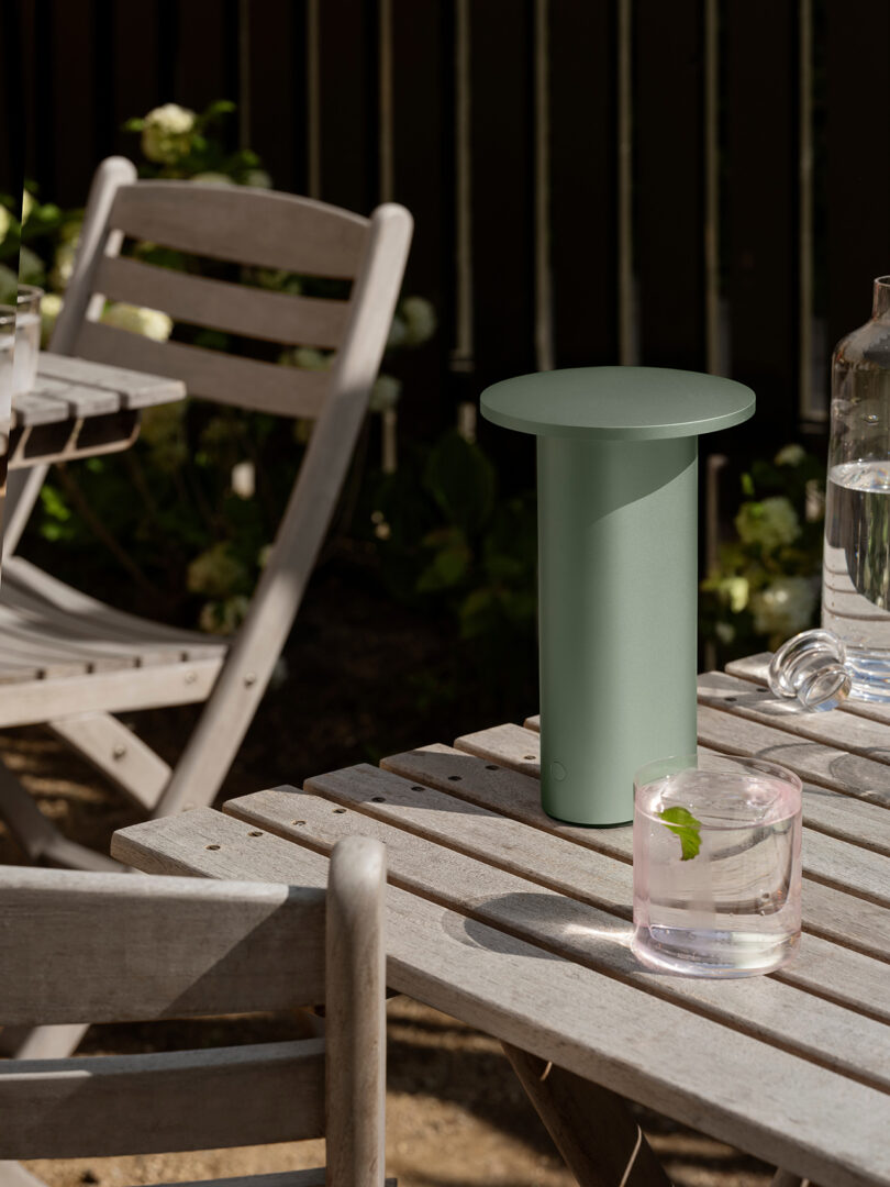 A matte green minimalist lamp on a wooden table surrounded by lush greenery.