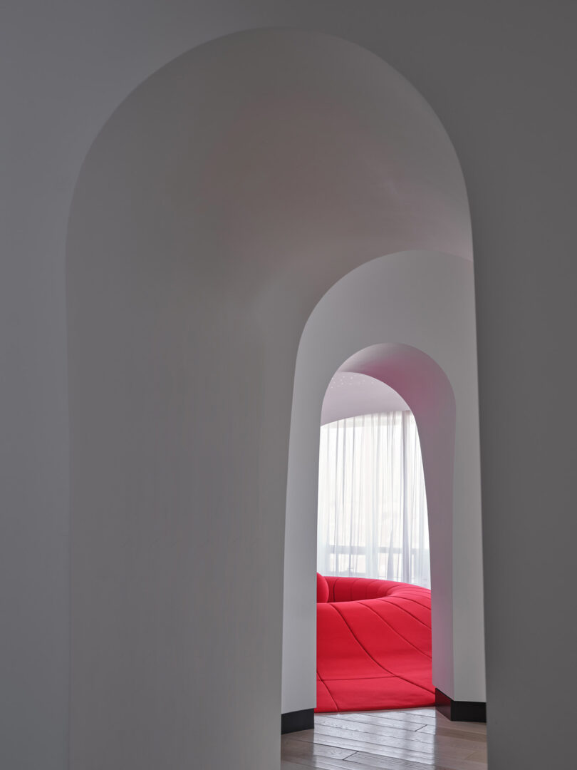 A smooth white arched doorway framing a view of a red modern sofa and sunlit curtains.