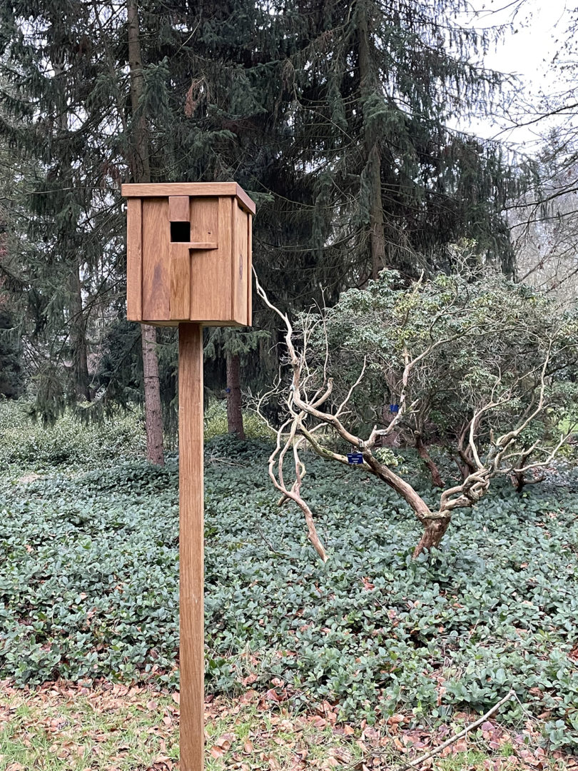 A modern wood bird nesting box mounted on a metal stake in a sunny yard.