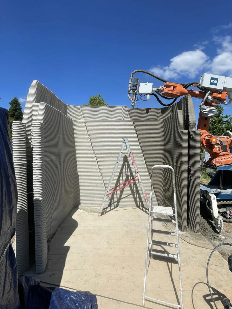 A robotic arm 3D printing the concrete layers of a building's wall on a construction site.