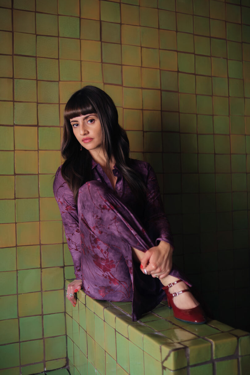 Maye Ruiz sitting on a green tiled ledge in a purple patterned outfit and red shoes.