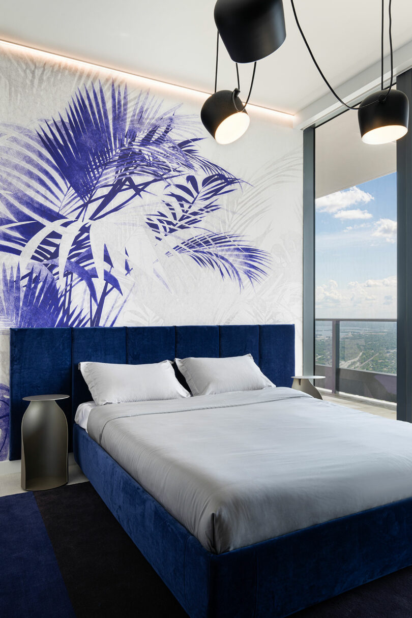 Bedroom with a blue velvet bed and a wall mural featuring blue palm leaves overlooking the city.