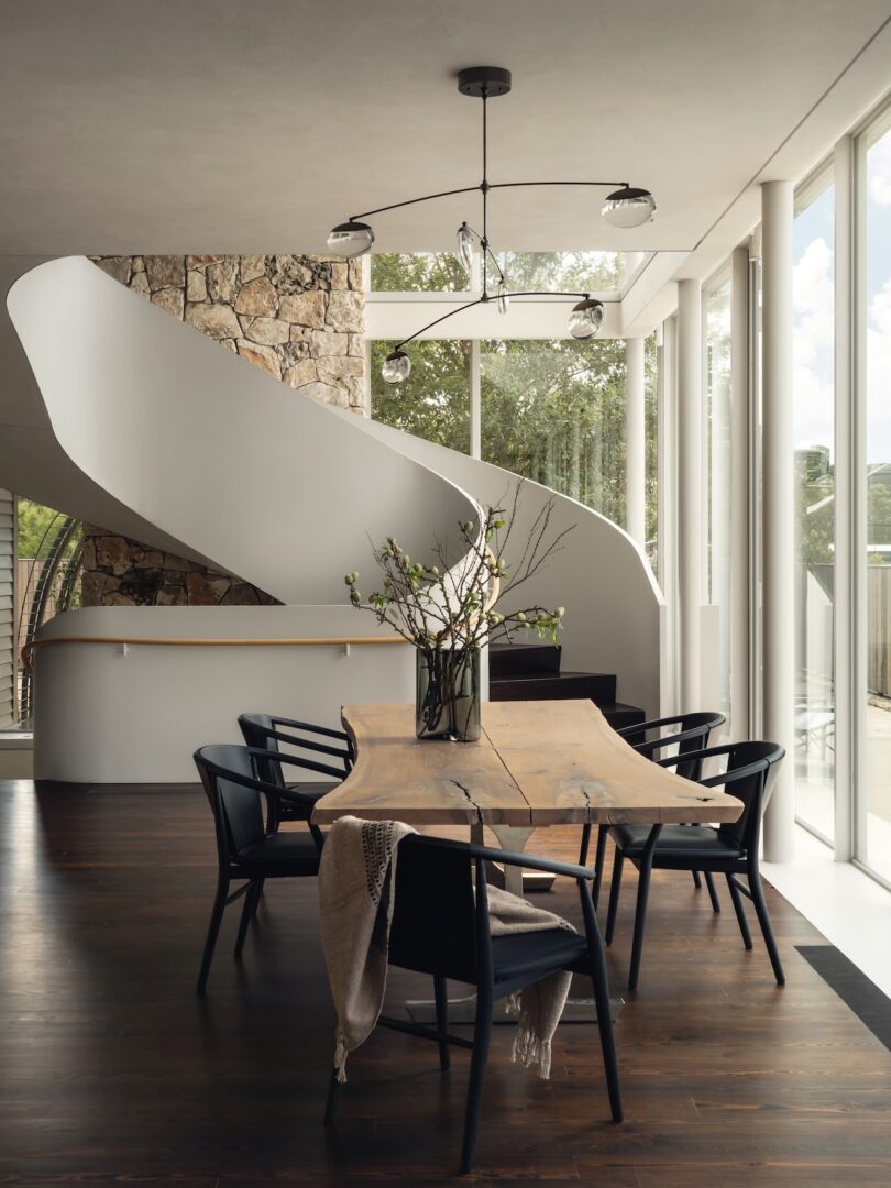 Modern dining room featuring a stone wall, minimalist wooden table, and a glimpse of a curved staircase.