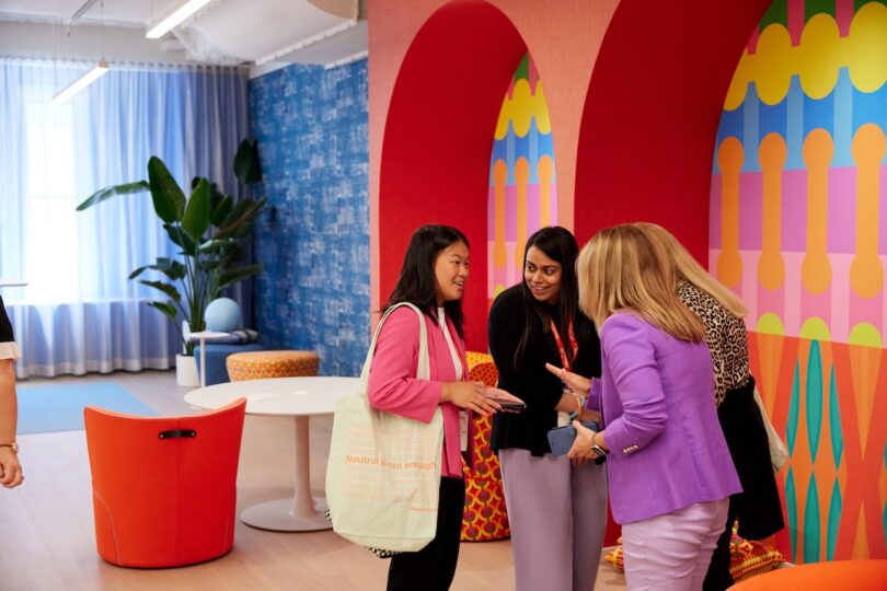 Four professional women talking in a bright, modern office space with bold patterns and greenery.