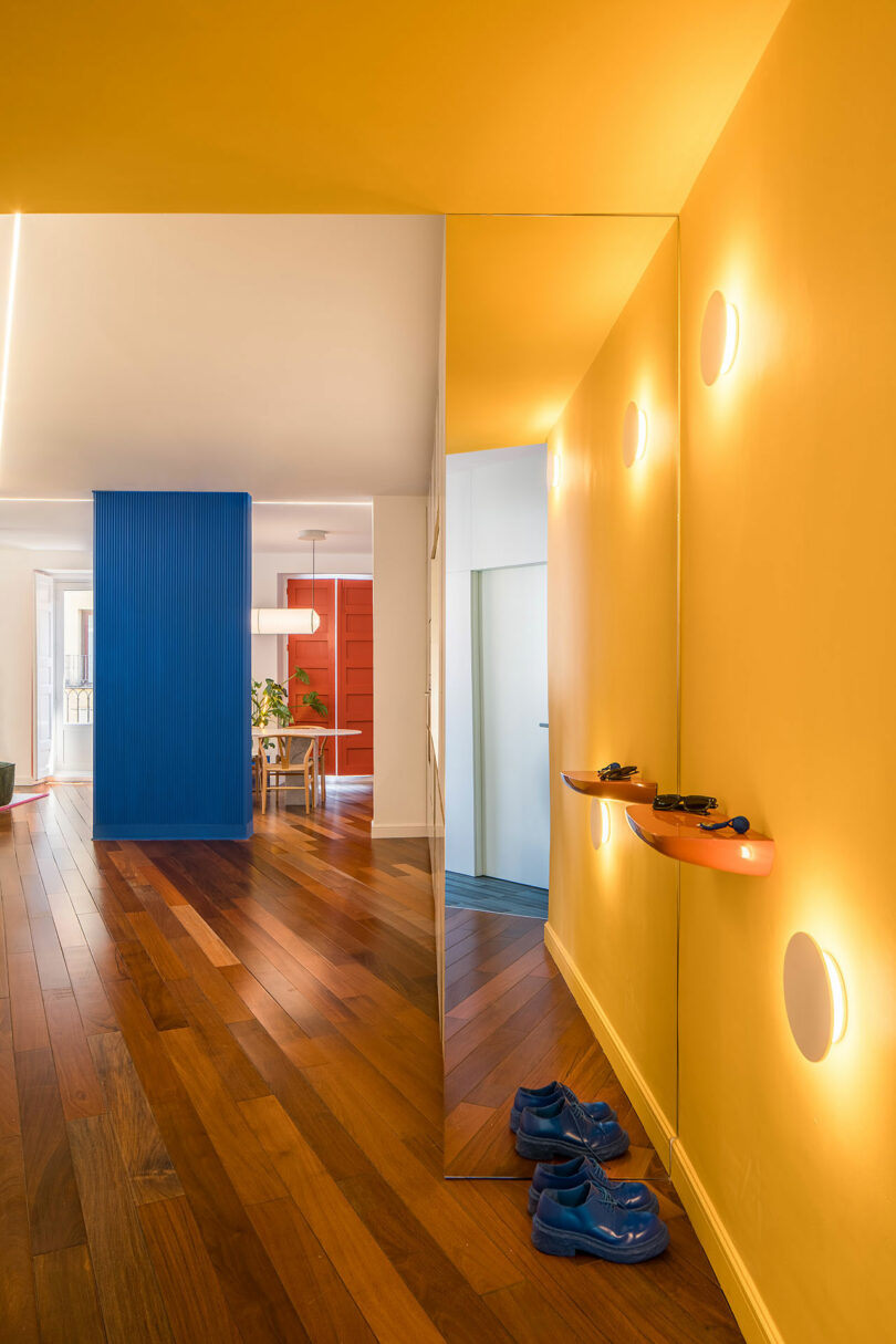 A yellow-walled hallway with a mirror and blue shoes, looking into a space with blue and red accent walls.