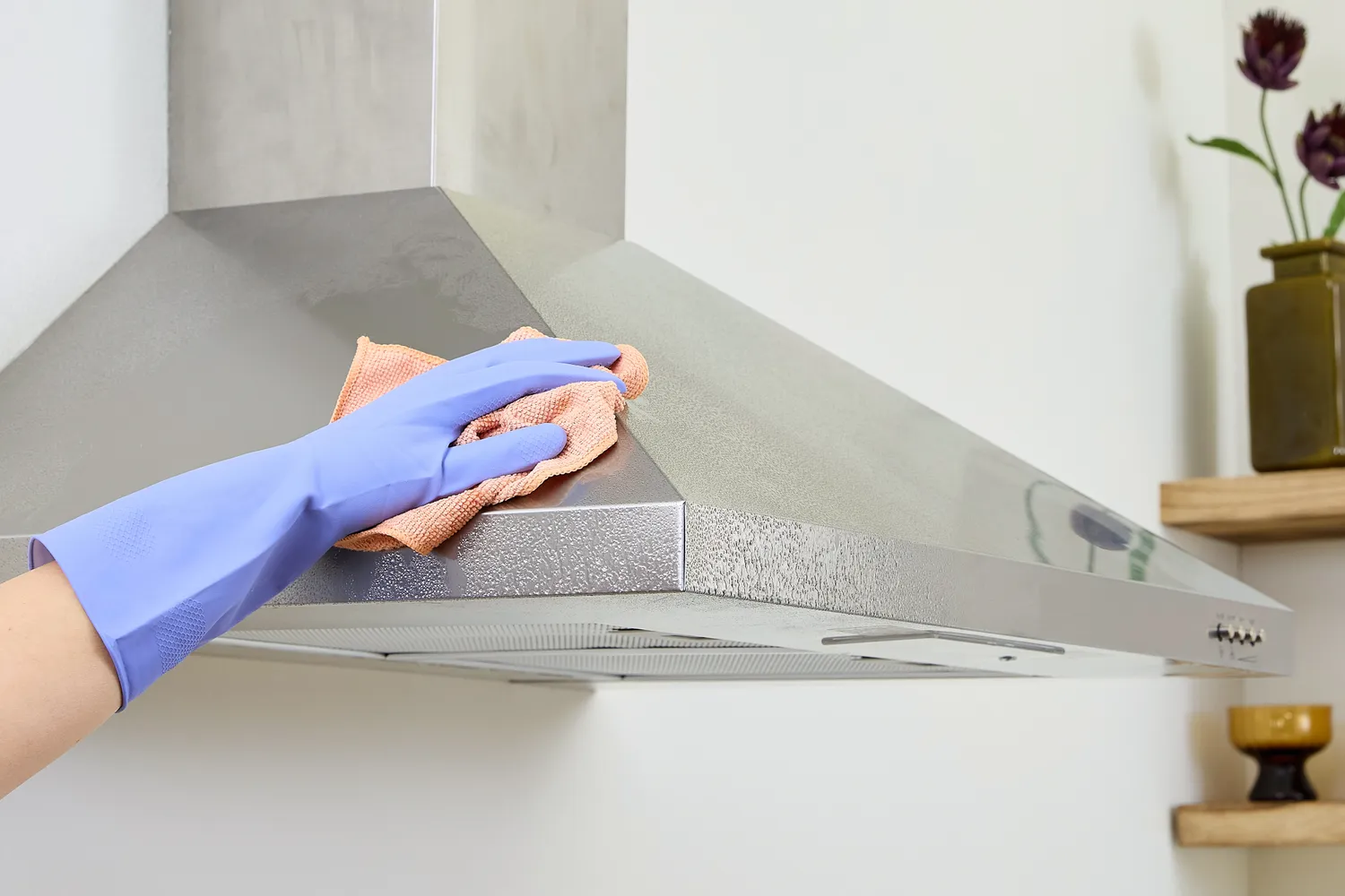 A person in gloves cleaning a stainless steel range hood in a contemporary kitchen.