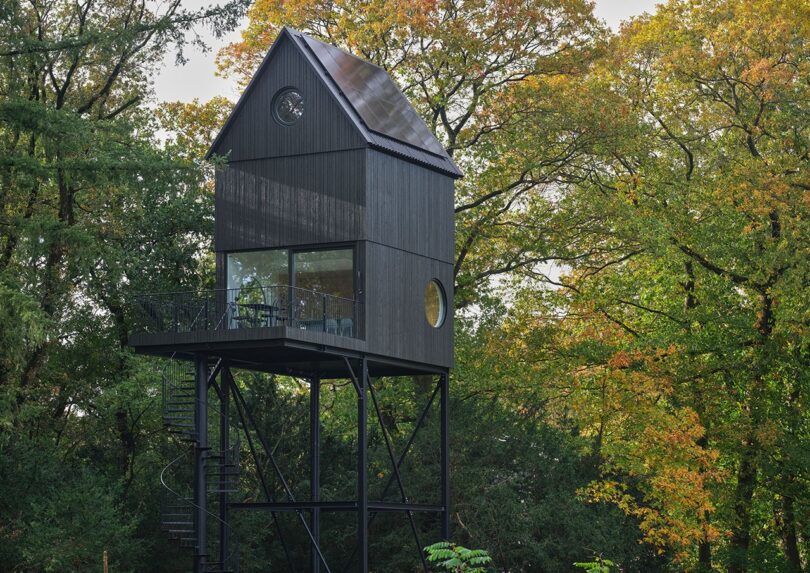 A modern black treehouse structure on stilts nestled among autumn trees.