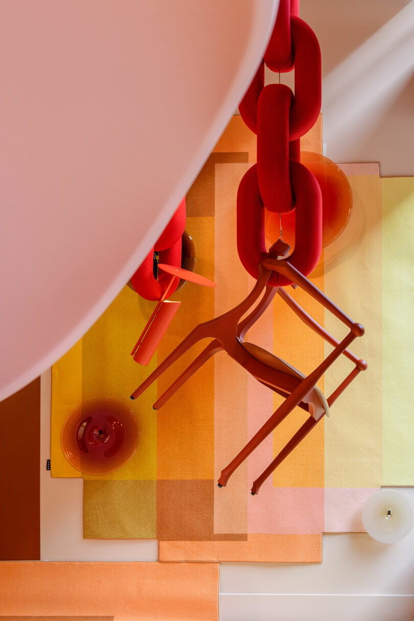 Top-down view of red Faye Toogood Roly Poly chairs suspended by oversized red links over colorful rugs.