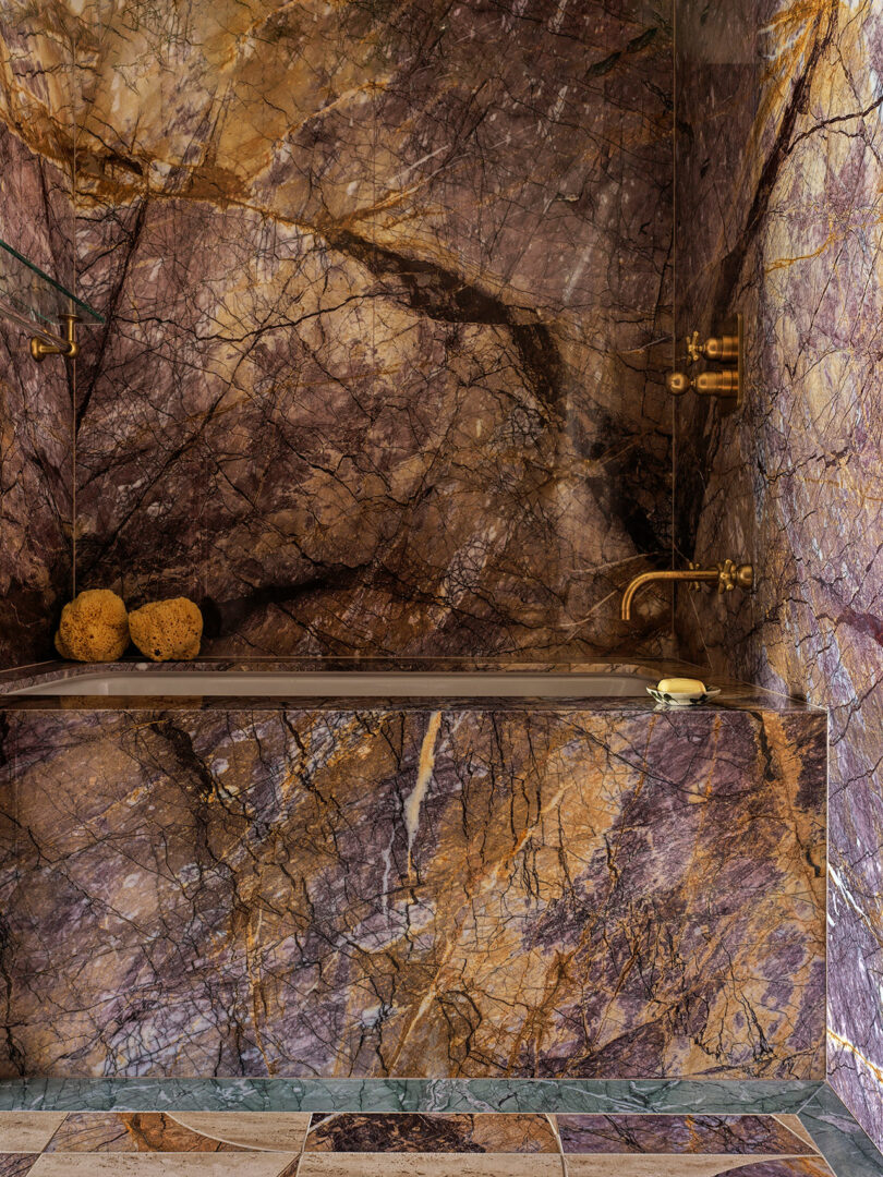 A luxury bathroom clad entirely in bookmatched multicolored marble with brass fixtures.