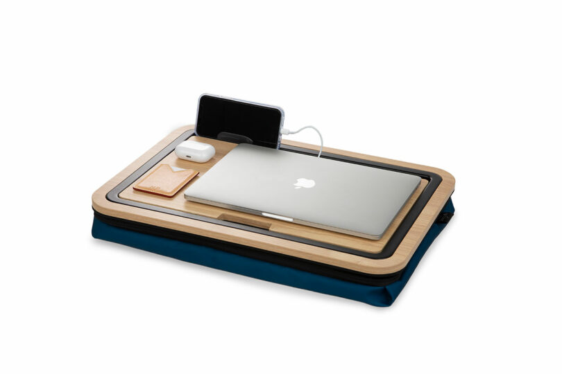 Close-up of a lap desk with a laptop, charging smartphone, and earbuds case.