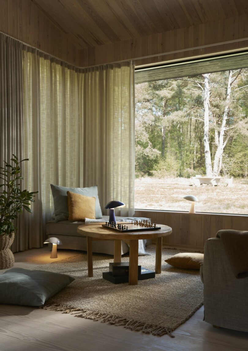 A cozy living room with a round wooden table, a chessboard, and a view of trees through a window.