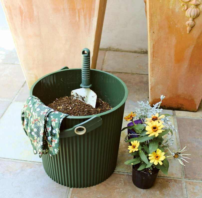 A green Hachiman bucket on a tiled floor filled with gardening soil and tools.