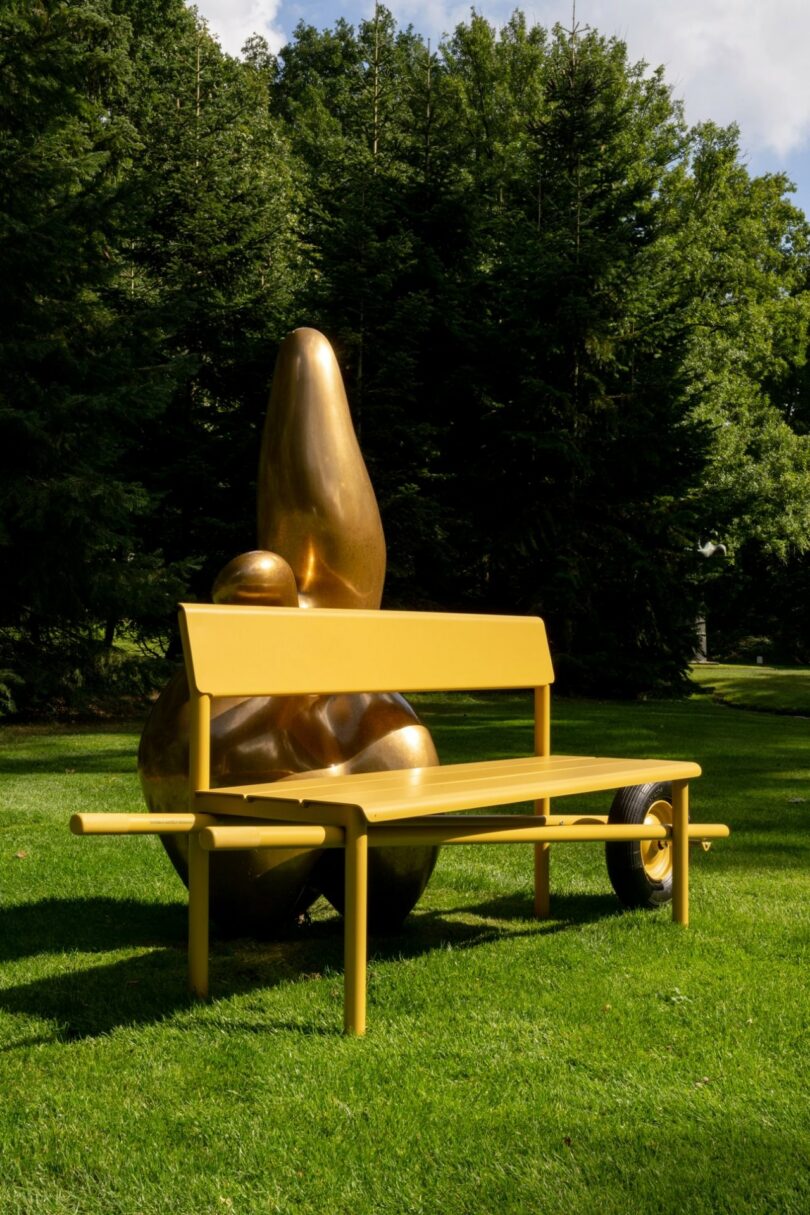 A yellow Wheelbench placed in front of a modern bronze sculpture in a wooded area.