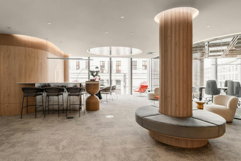 Modern office lounge with curved seating, a wooden social hub, and large windows overlooking the city.