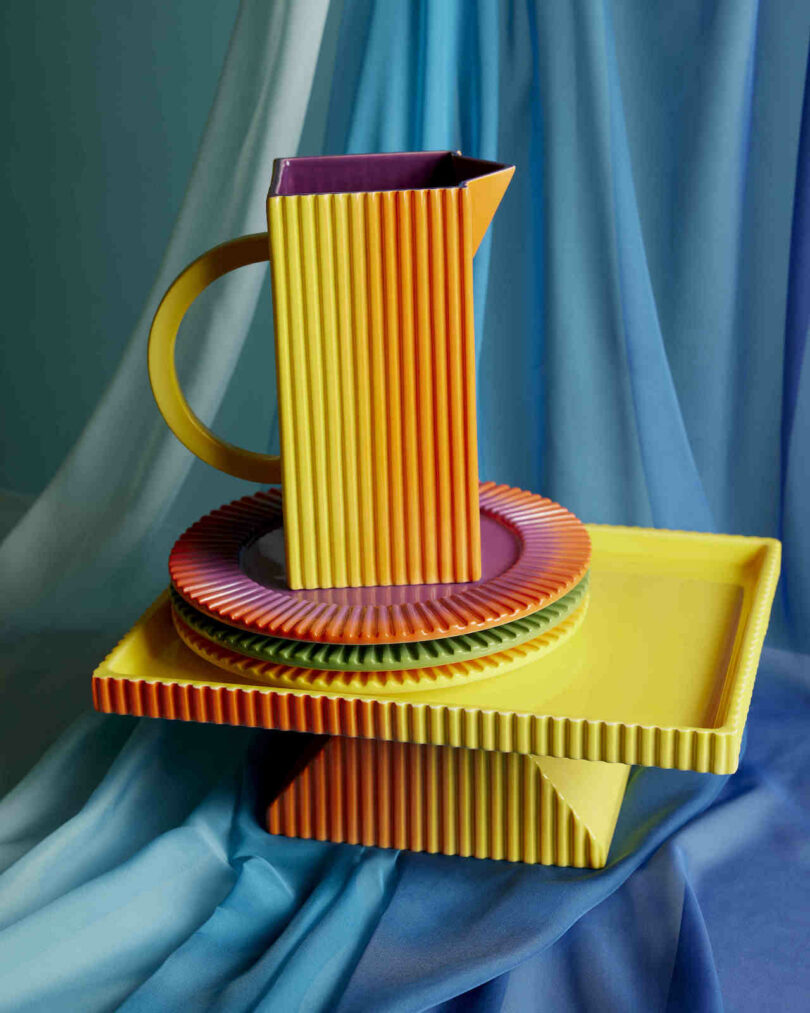 An arrangement of blue and orange ribbed ceramic jugs and plates on layered fabric.