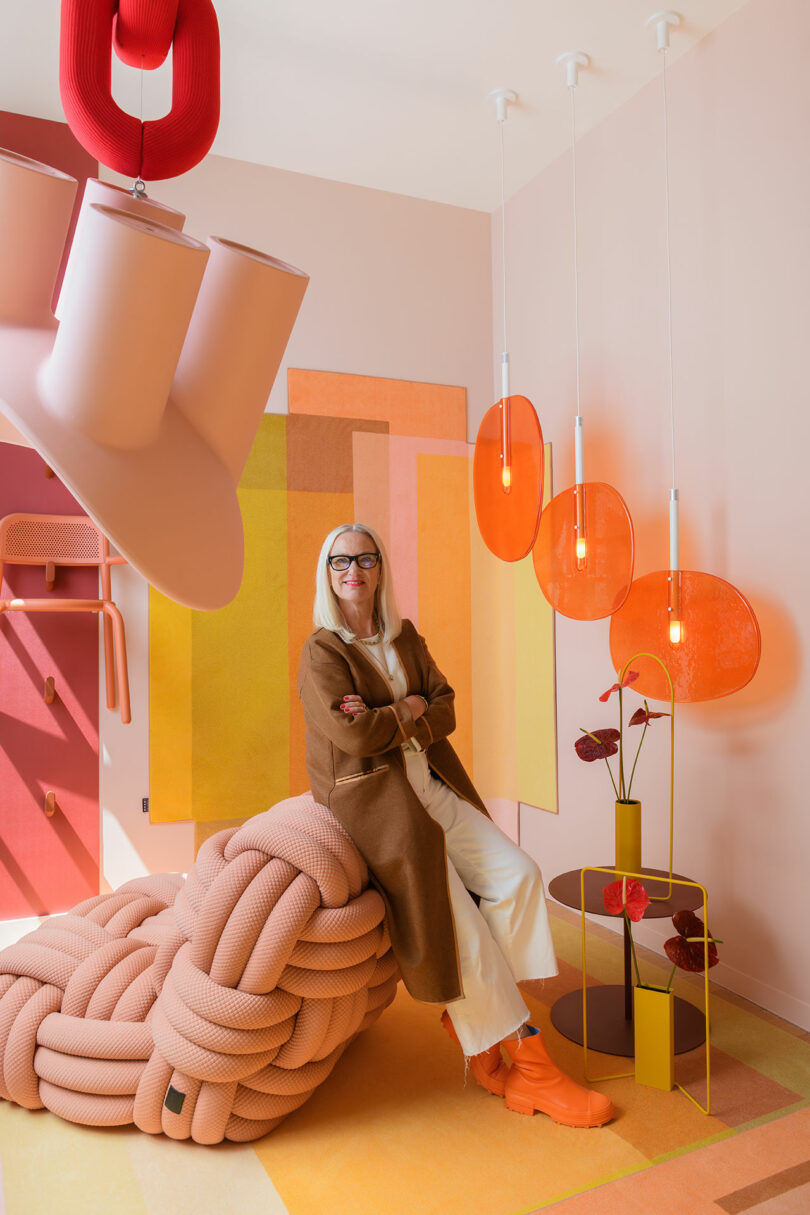 A woman sitting on a pink knotted chair in a room filled with orange lighting and geometric wall art.