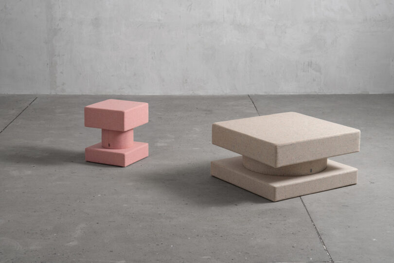 A pink and a beige geometric TAITŌ stool placed on a concrete floor against a textured gray wall.